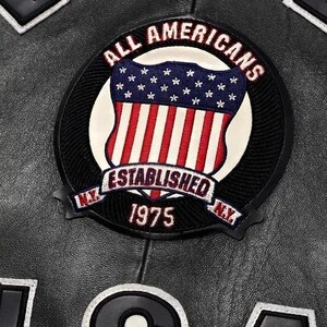 May include: Close-up of a black leather jacket with a circular patch. The patch features a shield design with the American flag and the words "ALL AMERICANS ESTABLISHED 1975 N.Y." in a circular design. Other patches are visible.