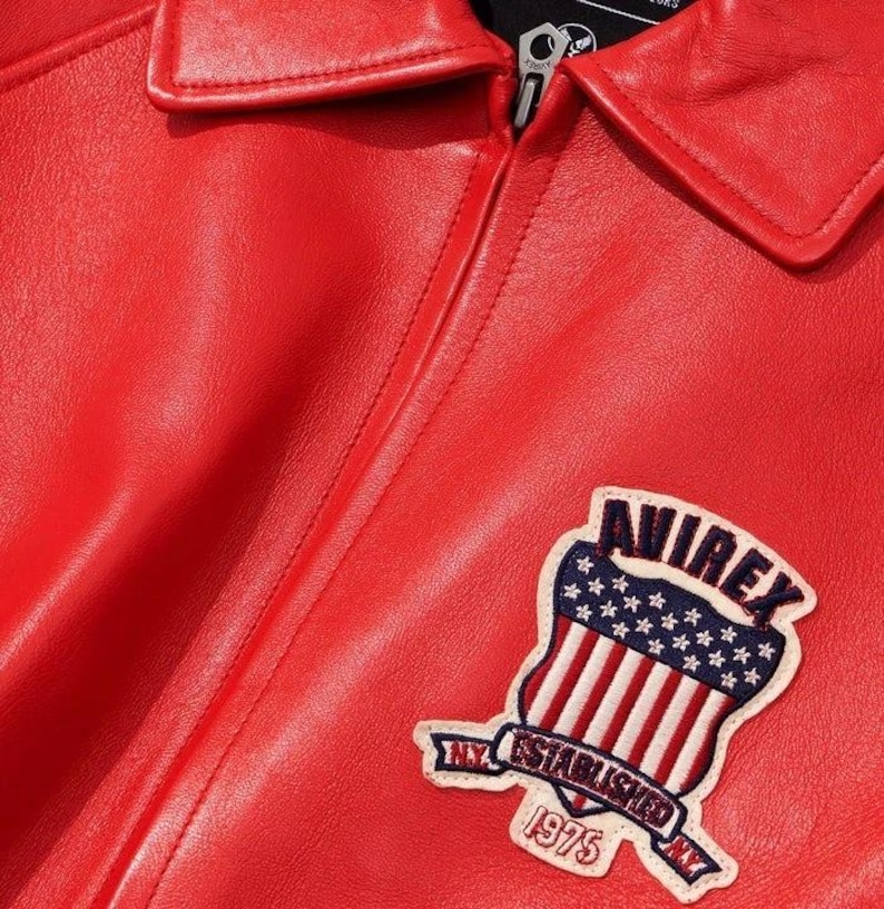 Peut inclure: Gros plan sur une veste en cuir rouge vif avec un col classique. La veste pr&eacute;sente un &eacute;cusson avec le texte "AVIREX" au-dessus d'un motif de bouclier avec le drapeau am&eacute;ricain et le texte "NY ESTABLISHED 1975".