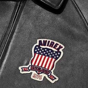 May include: Close-up of a black leather jacket with a decorative patch. The patch features the word "AVIREX" above a shield design with red and white stripes and blue stars. The text "ESTABLISHED NY 1975" is also visible.