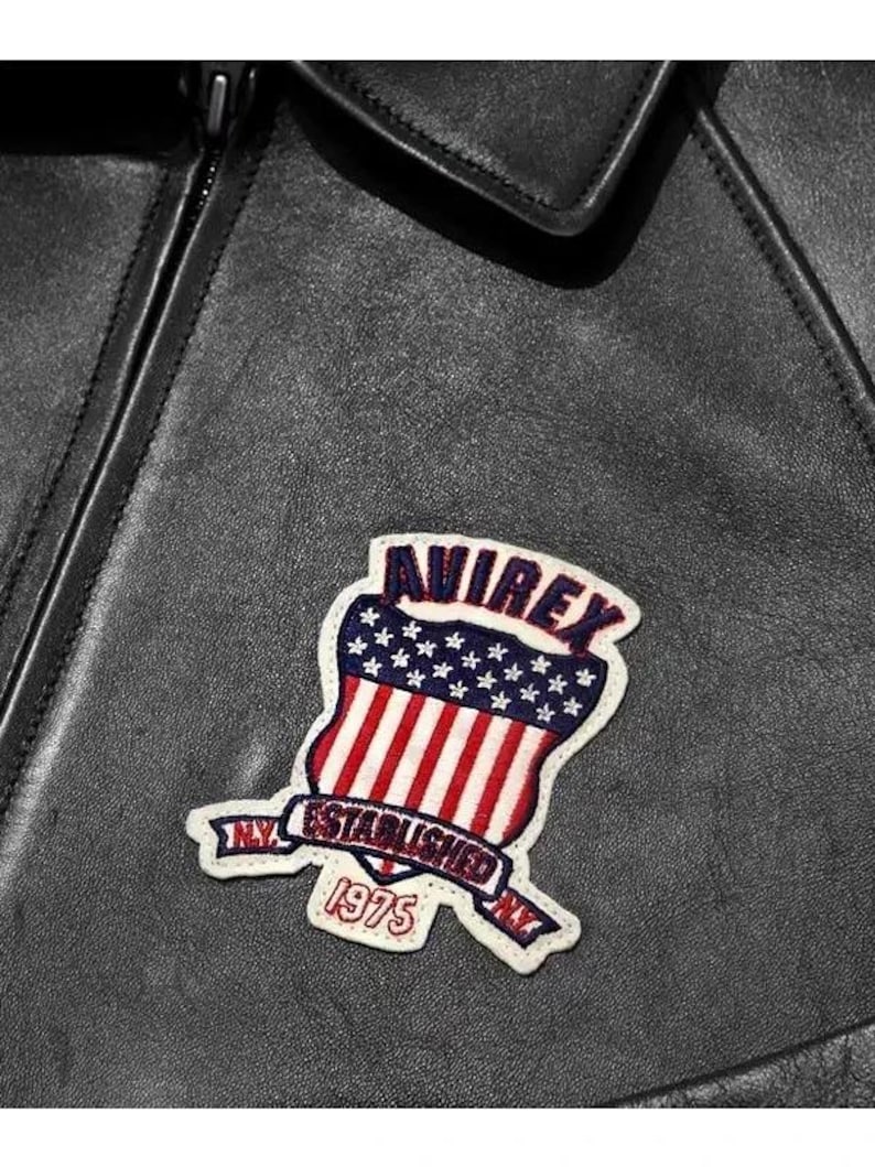 May include: Close-up of a black leather jacket with a decorative patch. The patch features the word "AVIREX" above a shield design with red and white stripes and blue stars. The text "ESTABLISHED NY 1975" is also visible.
