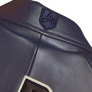 K&ouml;nnte beinhalten: Nahaufnahme einer marineblauen Lederjacke. Die Jacke hat ein marineblaues Emblem am oberen R&uuml;cken und einen wei&szlig;en und schwarzen Aufn&auml;her am unteren Rand. Das Leder hat eine strukturierte Optik.