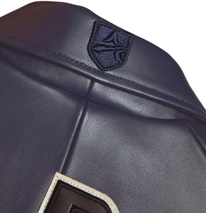 K&ouml;nnte beinhalten: Nahaufnahme einer marineblauen Lederjacke. Die Jacke hat ein marineblaues Emblem am oberen R&uuml;cken und einen wei&szlig;en und schwarzen Aufn&auml;her am unteren Rand. Das Leder hat eine strukturierte Optik.