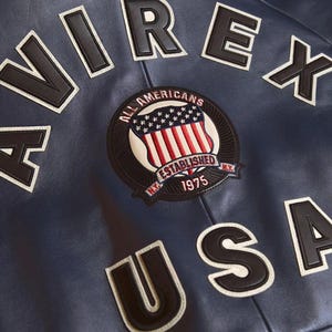 K&ouml;nnte beinhalten: Nahaufnahme einer marineblauen Lederjacke mit wei&szlig;en und schwarzen Buchstaben, die "AVIREX USA" buchstabieren. Ein kreisf&ouml;rmiger Aufn&auml;her zeigt einen Schild mit der amerikanischen Flagge und den Worten "ALL AMERICANS ESTABLISHED 1975".