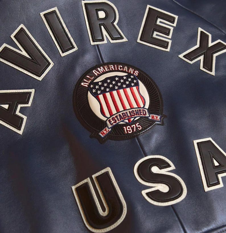 K&ouml;nnte beinhalten: Nahaufnahme einer marineblauen Lederjacke mit wei&szlig;en und schwarzen Buchstaben, die "AVIREX USA" buchstabieren. Ein kreisf&ouml;rmiger Aufn&auml;her zeigt einen Schild mit der amerikanischen Flagge und den Worten "ALL AMERICANS ESTABLISHED 1975".