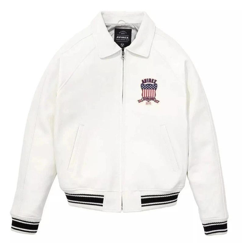 Puede incluir: Chaqueta blanca con cuello, cierre de cremallera y pu&ntilde;os y dobladillo a rayas blancas y negras. Presenta un logotipo en el pecho con el texto "AVIREX" y un dise&ntilde;o de bandera americana. Un estilo cl&aacute;sico de chaqueta bomber.