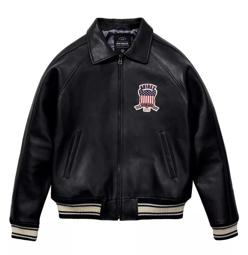 May include: Black leather jacket with a collared neckline and a zip-up front. The jacket features a patch on the chest with the text "AVIREX" and a design incorporating the American flag. The cuffs and hem have a striped design.