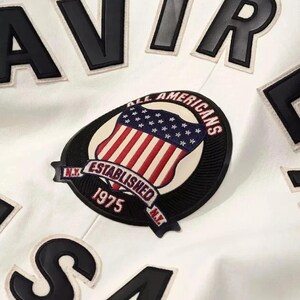 Puede incluir: Primer plano de una chaqueta blanca con letras y parches negros. La chaqueta presenta un parche con un dise&ntilde;o de escudo, la bandera estadounidense y el texto "ALL AMERICANS ESTABLISHED 1975 N.Y."
