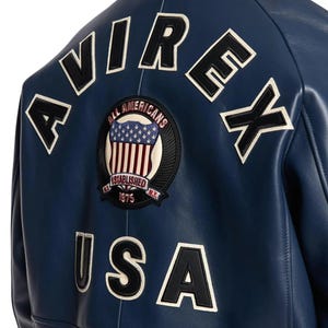 K&ouml;nnte beinhalten: Nahaufnahme einer marineblauen Lederjacke. Auf der R&uuml;ckseite der Jacke befinden sich wei&szlig;e und schwarze Buchstaben, die "AVIREX" und "USA" buchstabieren. Ein kreisf&ouml;rmiger Aufn&auml;her zeigt ein amerikanisches Flaggen-Design mit den Worten "ALL AMERICANS" und "ESTABLISHED 1975 N.Y."
