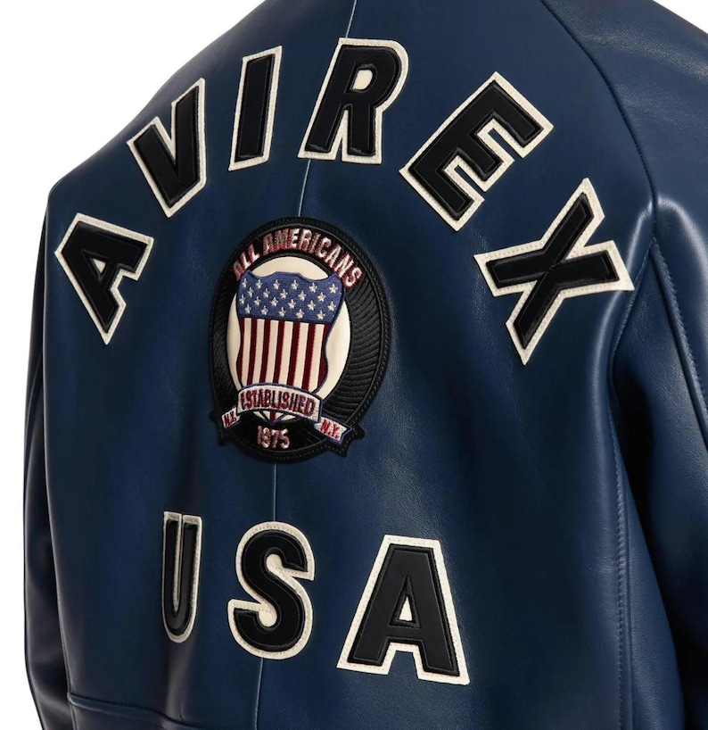 K&ouml;nnte beinhalten: Nahaufnahme einer marineblauen Lederjacke. Auf der R&uuml;ckseite der Jacke befinden sich wei&szlig;e und schwarze Buchstaben, die "AVIREX" und "USA" buchstabieren. Ein kreisf&ouml;rmiger Aufn&auml;her zeigt ein amerikanisches Flaggen-Design mit den Worten "ALL AMERICANS" und "ESTABLISHED 1975 N.Y."