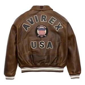 Op de afbeelding: Bruine leren jas met het woord "AVIREX" over de schouders en "USA" aan de onderkant. Een cirkelvormige patch met de Amerikaanse vlag en de woorden "ALL AMERICANS ESTABLISHED 1975" is gecentreerd. De jas heeft wit en bruin gestreepte manchetten en zoom.