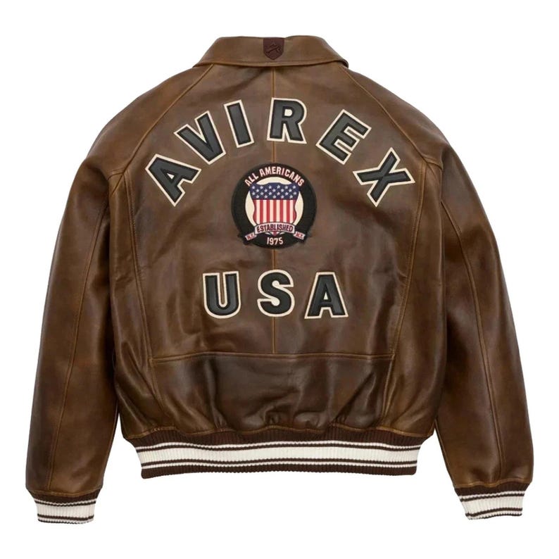 Op de afbeelding: Bruine leren jas met het woord "AVIREX" over de schouders en "USA" aan de onderkant. Een cirkelvormige patch met de Amerikaanse vlag en de woorden "ALL AMERICANS ESTABLISHED 1975" is gecentreerd. De jas heeft wit en bruin gestreepte manchetten en zoom.