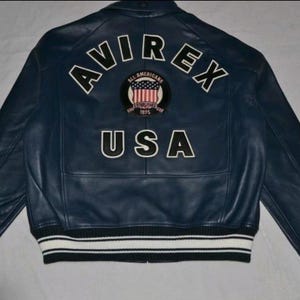 K&ouml;nnte beinhalten: Marineblaue Lederjacke mit wei&szlig;-schwarz gestreiften B&uuml;ndchen und Saum. Auf dem R&uuml;cken steht das Wort "AVIREX" &uuml;ber einem runden Aufn&auml;her und dem Wort "USA".