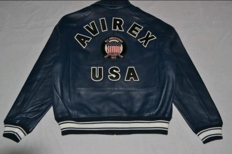 K&ouml;nnte beinhalten: Marineblaue Lederjacke mit wei&szlig;-schwarz gestreiften B&uuml;ndchen und Saum. Auf dem R&uuml;cken steht das Wort "AVIREX" &uuml;ber einem runden Aufn&auml;her und dem Wort "USA".