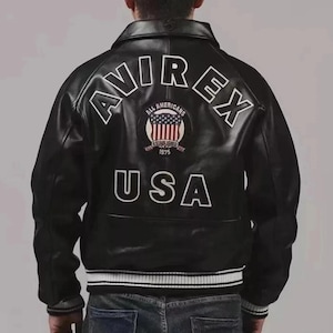 May include: Black leather jacket with white lettering that reads "AVIREX" and "USA". The back of the jacket features a patch with the words "ALL AMERICANS" and "ESTABLISHED 1975". The jacket has a ribbed collar and cuffs.