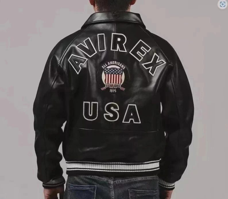May include: Black leather jacket with white lettering that reads "AVIREX" and "USA". The back of the jacket features a patch with the words "ALL AMERICANS" and "ESTABLISHED 1975". The jacket has a ribbed collar and cuffs.