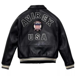 May include: Black leather jacket with white embroidered lettering and a patch featuring the American flag. The jacket has a classic bomber style with a ribbed collar, cuffs, and hem. The text on the back reads "AVIREX" and "USA".