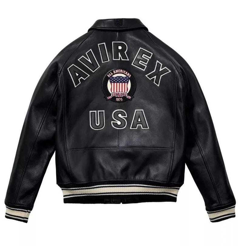 May include: Black leather jacket with white embroidered lettering and a patch featuring the American flag. The jacket has a classic bomber style with a ribbed collar, cuffs, and hem. The text on the back reads "AVIREX" and "USA".