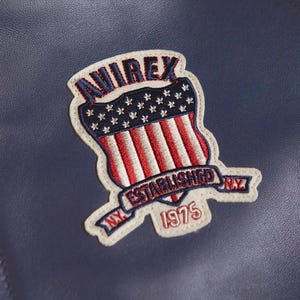 K&ouml;nnte beinhalten: Nahaufnahme einer marineblauen Lederjacke mit einem aufgen&auml;hten Aufn&auml;her. Der Aufn&auml;her zeigt das Wort "AVIREX" in Rot und Blau, einen Schild mit roten und wei&szlig;en Streifen und Sternen sowie den Text "ESTABLISHED N.Y. 1975".