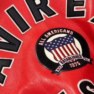 Peut inclure: Gros plan sur une veste en cuir rouge avec des lettres noires et blanches &eacute;pelant "AIRE" et "RE". Un &eacute;cusson circulaire pr&eacute;sente un drapeau am&eacute;ricain avec le texte "ALL AMERICANS ESTABLISHED 1975 N.Y."