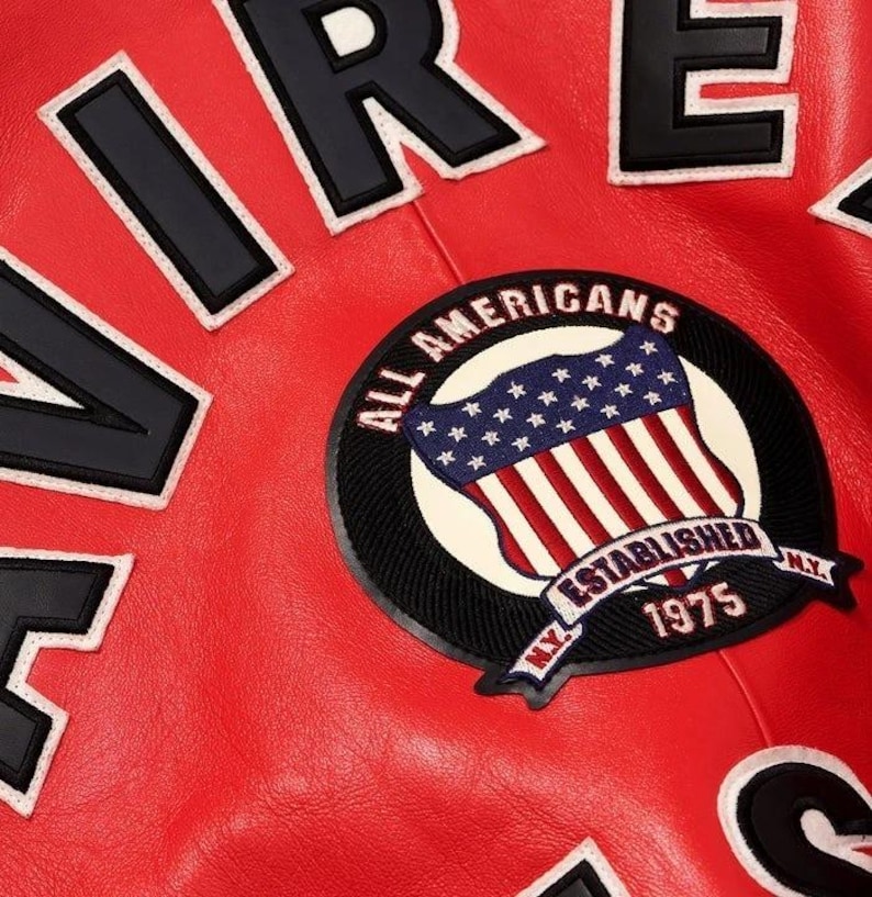 Peut inclure: Gros plan sur une veste en cuir rouge avec des lettres noires et blanches &eacute;pelant "AIRE" et "RE". Un &eacute;cusson circulaire pr&eacute;sente un drapeau am&eacute;ricain avec le texte "ALL AMERICANS ESTABLISHED 1975 N.Y."