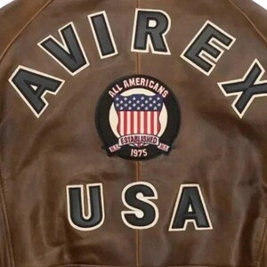 Op de afbeelding: Bruine leren jas met het woord "AVIREX" in witte letters met zwarte contouren. Een cirkelvormige patch toont "ALL AMERICANS ESTABLISHED 1975 NY" met een ontwerp van de Amerikaanse vlag. Het woord "USA" staat eronder.