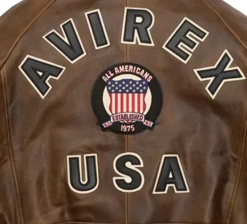 Op de afbeelding: Bruine leren jas met het woord "AVIREX" in witte letters met zwarte contouren. Een cirkelvormige patch toont "ALL AMERICANS ESTABLISHED 1975 NY" met een ontwerp van de Amerikaanse vlag. Het woord "USA" staat eronder.