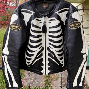 May include: Black leather jacket with a white skeleton design. Features a full zip closure, two patches with the "Vanson Leathers" logo, and a classic biker jacket silhouette. The jacket is hanging on a wooden hanger.
