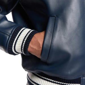 K&ouml;nnte beinhalten: Nahaufnahme einer marineblauen Lederjacke mit geripptem Strickb&uuml;ndchen. Das B&uuml;ndchen hat wei&szlig;e Streifen auf marineblauem Hintergrund. Die Jacke hat eine Tasche und eine glatte, gl&auml;nzende Textur.