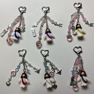 May include: Ten colourful keychains featuring a small figurine of Jesus wearing a white robe with pink, purple, or red trim. Each keychain has a variety of charms, including strawberries, hearts, music notes, and mobile phones.