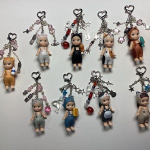 May include: A collection of small, decorative keychains featuring miniature doll figures in various animal costumes. Each keychain has a heart-shaped clasp and is adorned with beads, charms, and dangling embellishments.