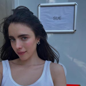 May include: A woman with long dark hair wearing a white tank top and a star-shaped earring. A silver frame with the word "SUE" is visible in the background.