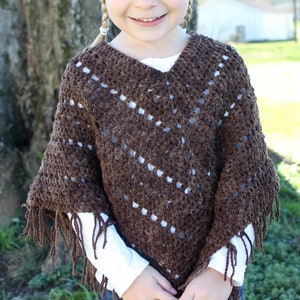 May include: A young girl wearing a brown crocheted poncho with fringe. The poncho has a V-neck and a diamond pattern.