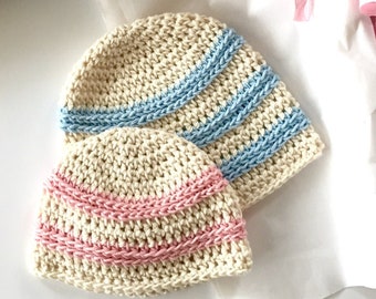May include: Two crocheted baby hats, one with blue stripes and one with pink stripes, on a white surface.