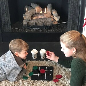 May include: Two children play a game of tic-tac-toe on a black and white crocheted board with red and green felt game pieces. The children are sitting on a fluffy rug in front of a fireplace.