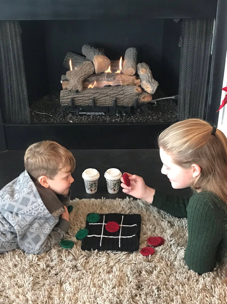 May include: Two children play a game of tic-tac-toe on a black and white crocheted board with red and green felt game pieces. The children are sitting on a fluffy rug in front of a fireplace.