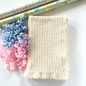 May include: A cream-colored crocheted baby blanket with a scalloped edge. The blanket is surrounded by blue and pink curling ribbon.