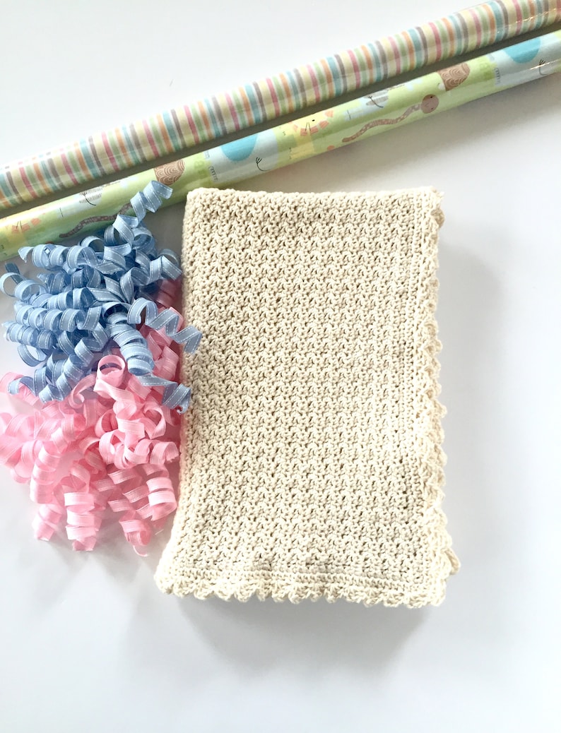 May include: A cream-colored crocheted baby blanket with a scalloped edge. The blanket is surrounded by blue and pink curling ribbon.