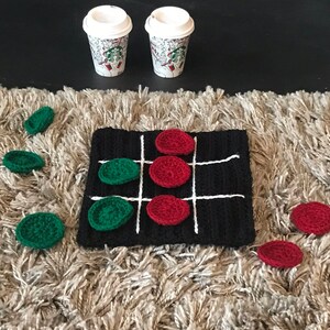 May include: A black and white crocheted tic-tac-toe board with red and green crocheted game pieces. There are two coffee cups on a black surface above the board.