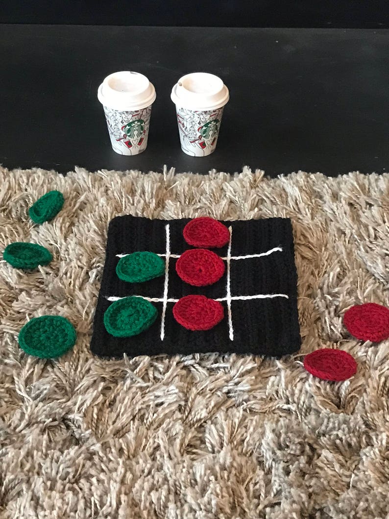 May include: A black and white crocheted tic-tac-toe board with red and green crocheted game pieces. There are two coffee cups on a black surface above the board.