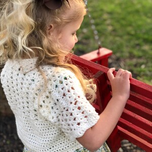 May include: A young girl with long blonde hair wearing a white crocheted top and a floral skirt. She is holding onto a red wooden swing set.