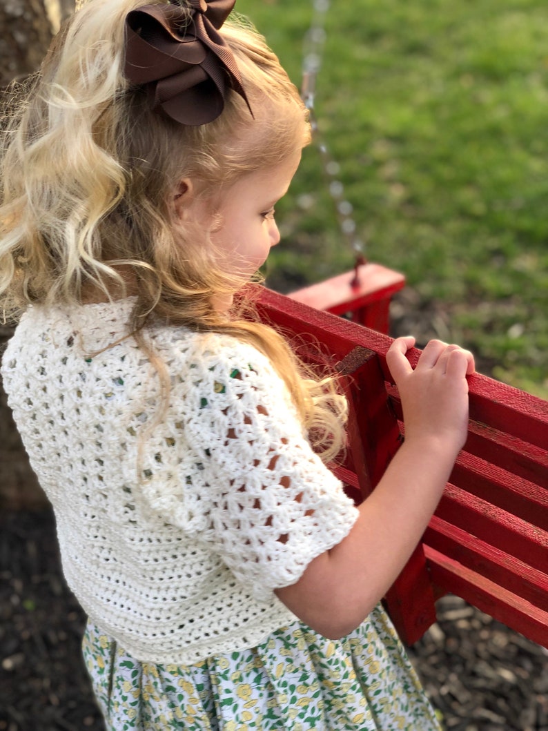 May include: A young girl with long blonde hair wearing a white crocheted top and a floral skirt. She is holding onto a red wooden swing set.