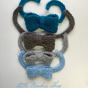 Puede incluir: Cuatro pajaritas de ganchillo en diferentes colores: azul verdoso, gris, gris claro y azul claro. Las pajaritas están apiladas, con la pajarita azul verdosa en la parte superior. Todas las pajaritas están hechas con un patrón de punto sencillo y tienen un aspecto ligeramente texturizado. El texto "Little Monkey Shop" está escrito en cursiva azul en la parte inferior de la imagen.