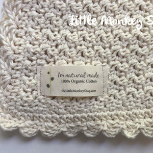 May include: A close-up of a cream-colored crocheted baby blanket with a scalloped edge. The blanket has a small label that reads "I'm natural made 100% Organic Cotton thelittlemonkeyshop.com".
