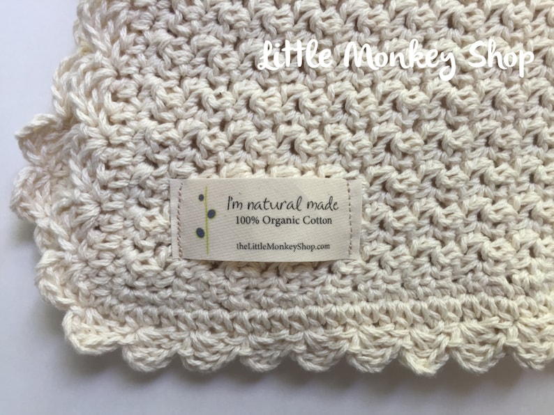 May include: A close-up of a cream-colored crocheted baby blanket with a scalloped edge. The blanket has a small label that reads "I'm natural made 100% Organic Cotton thelittlemonkeyshop.com".