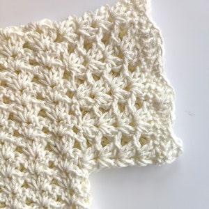 May include: Close-up of a white crocheted garment with a scalloped edge. The fabric is made with a delicate stitch pattern.