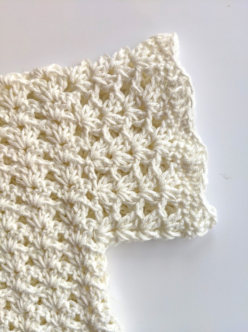 May include: Close-up of a white crocheted garment with a scalloped edge. The fabric is made with a delicate stitch pattern.