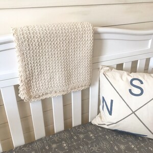 May include: A white crocheted baby blanket with a scalloped edge hangs on a white crib. The crib has a gray and white patterned sheet and a white pillow with a blue compass design and the letters "N" and "S" on it.