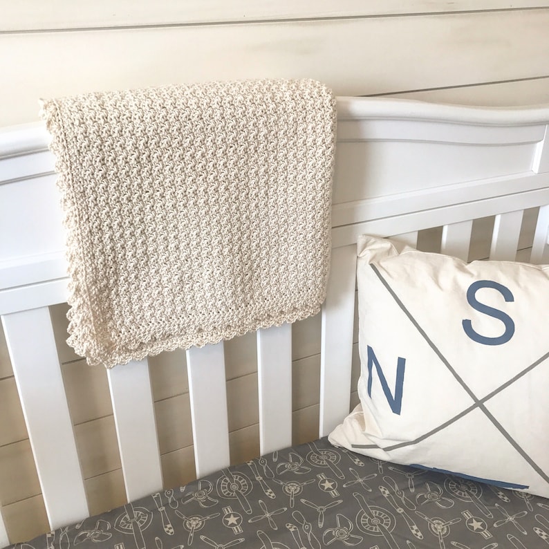 May include: A white crocheted baby blanket with a scalloped edge hangs on a white crib. The crib has a gray and white patterned sheet and a white pillow with a blue compass design and the letters "N" and "S" on it.