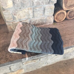May include: A crocheted blanket with a wavy pattern in shades of pink, brown, blue, and gray. The blanket is folded and lying on a stone surface.