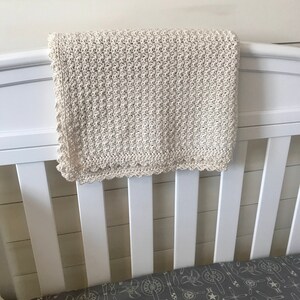 May include: A white crocheted baby blanket with a scalloped edge hangs on a white crib. The crib has a gray and white patterned sheet with a propeller design.